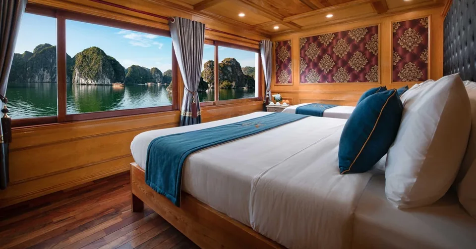 A wooden-panelled cruise cabin with a double bed and windows framing limestone karsts